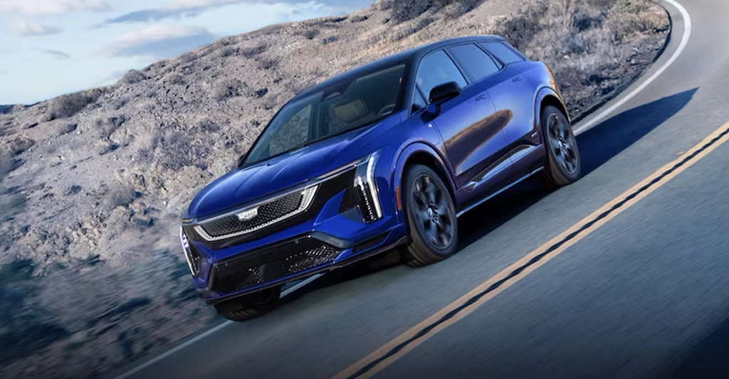 A vibrant, eye-level shot captures a blue Cadillac Lyriq SUV in motion on a winding road, set against a backdrop of a rocky mountain.