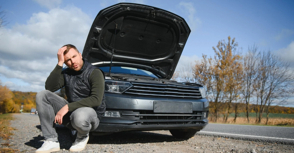 Frustrated man crouching beside a gray sedan with the hood open on a sunny roadside in autumn.