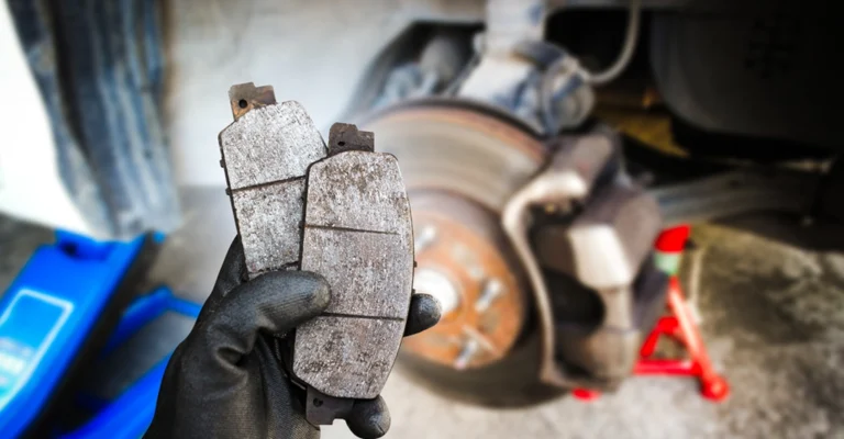 Close-up shot of a gloved hand holding a pair of used brake pads in front of a car's brake assembly.