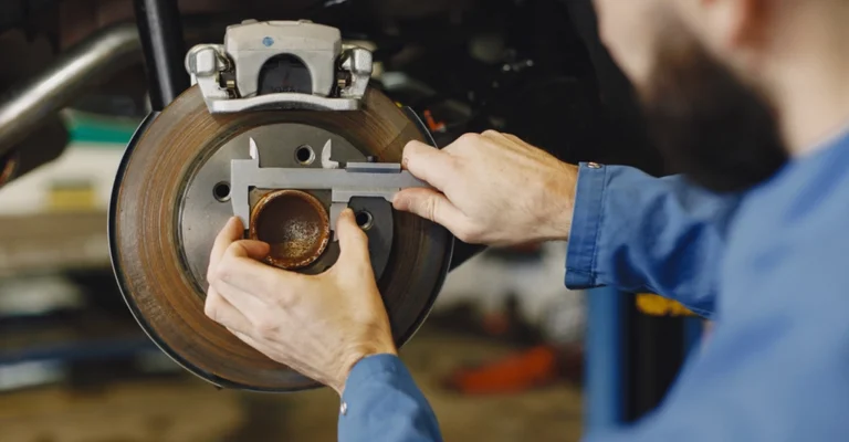 Caucasian male, around 35 years old, with a beard, wearing a blue uniform, using a caliper to measure a rusty car brake rotor.
