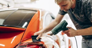 a fair-skinned, blonde-haired man, around 28 years old, meticulously applying a protective film to the rear of an orange car.