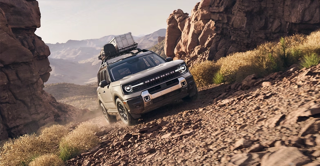 A tan Ford Bronco Sport is angled down a rocky, dusty slope, with a mountain range visible in the background.