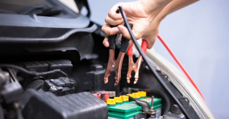 A close-up shot shows a light-skinned person holding jumper cables over a car battery in an engine compartment.