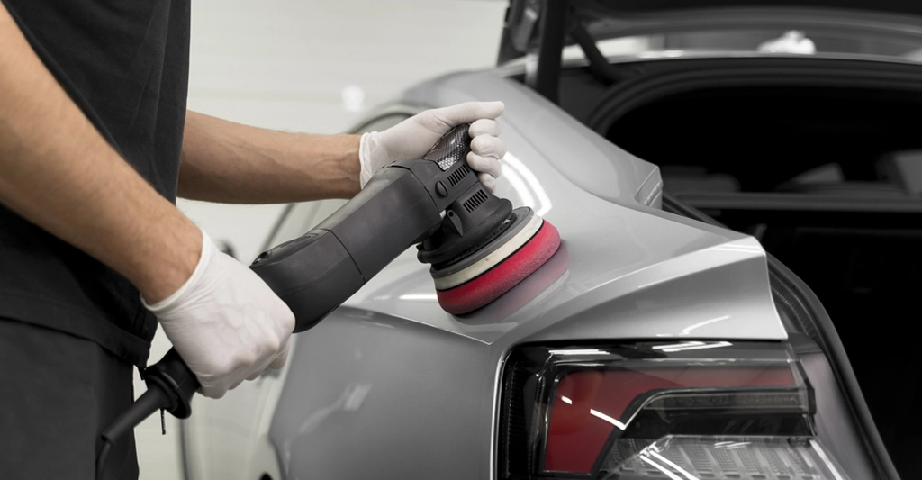 a light-skinned man, around 30 years old, wearing white gloves and a black t-shirt, polishing a silver car with a black and red polishing machine.