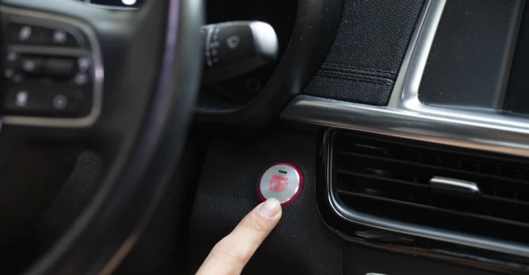 Close-up of a light-skinned hand pressing a silver engine start/stop button with a red ring and "ENGINE START STOP" text, set in a black textured dashboard with silver trim and nearby air vents; part of the steering wheel with controls is visible to the left under soft lighting.