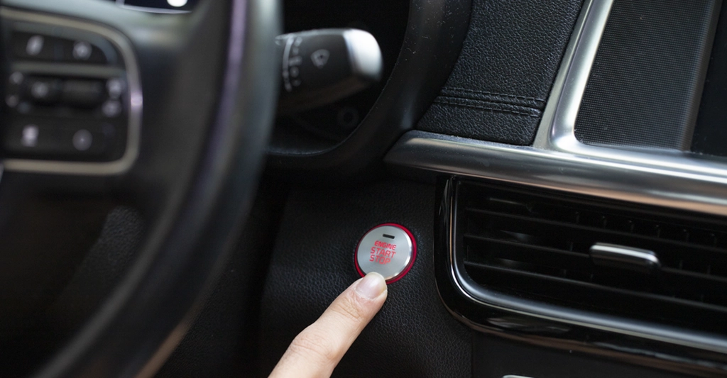 Close-up of a light-skinned hand pressing a silver engine start/stop button with a red ring and "ENGINE START STOP" text, set in a black textured dashboard with silver trim and nearby air vents; part of the steering wheel with controls is visible to the left under soft lighting.