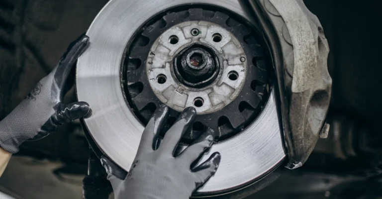 Close-up of a car's brake disc and caliper being inspected by gloved hands.
