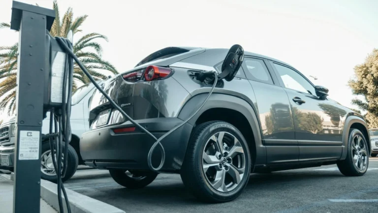 Gray Mazda MX-30 electric car charging at a station, plugged in with a black cable; rear and side profile visible, parked in a lot with palm trees and an overcast sky in the background.