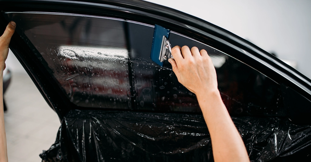 A close-up shot captures the process of tinting a car window, with a fair-skinned person's hands visible.