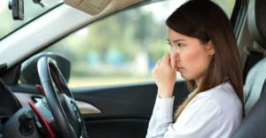 A medium shot depicts an Asian woman, around 25 years old, inside a car, pinching her nose with her right hand.