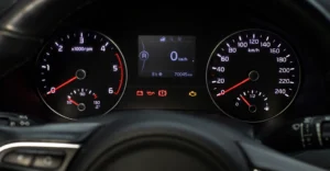 Close-up shot of a car dashboard, featuring two circular gauges on either side of a digital display. The left gauge measures RPM, with a red needle pointing to a low value, and a smaller gauge below indicating temperature