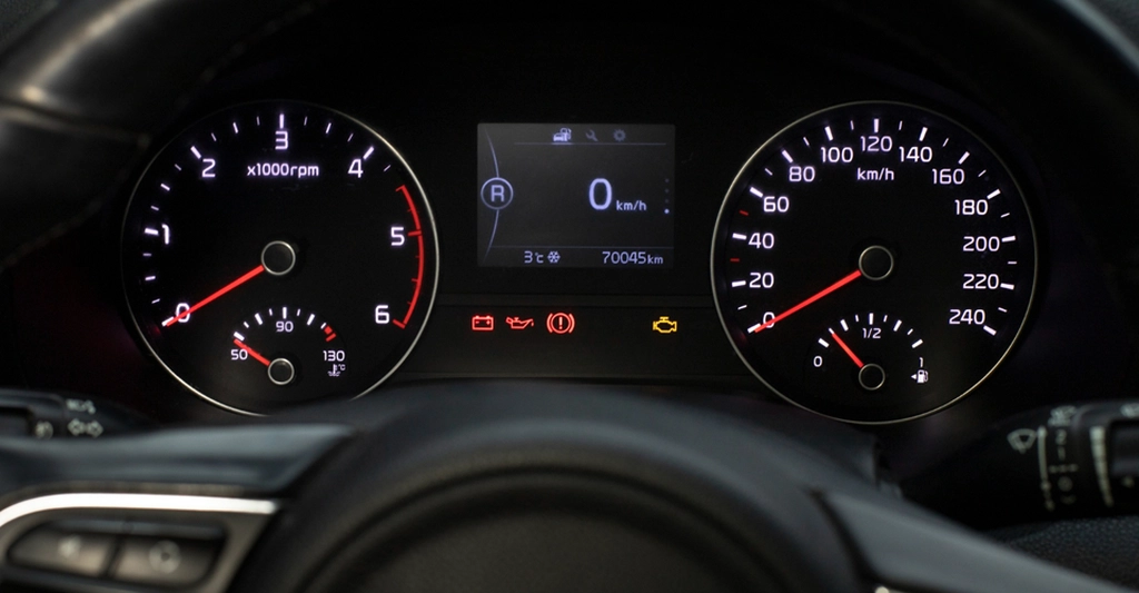 Close-up shot of a car dashboard, featuring two circular gauges on either side of a digital display. The left gauge measures RPM, with a red needle pointing to a low value, and a smaller gauge below indicating temperature