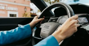 A close-up shot from inside a car shows a woman's hands on the steering wheel