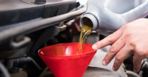 Close-up shot of a hand pouring motor oil from a gray plastic bottle into a red funnel, with the engine of a car visible in the background.