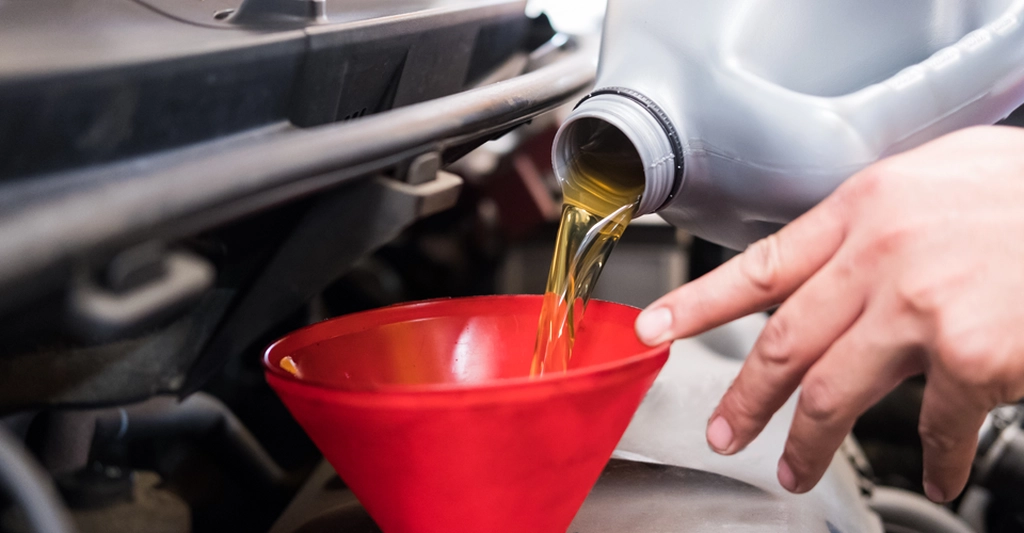 Close-up shot of a hand pouring motor oil from a gray plastic bottle into a red funnel, with the engine of a car visible in the background.