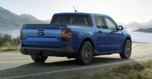 Blue 2025 Ford Maverick Hybrid Lariat pickup truck navigating a curved road beside a serene lake and mountains. The bright, sunny day adds a sense of adventure and freedom.
