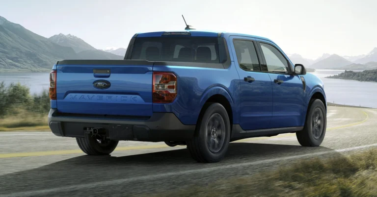 Blue 2025 Ford Maverick Hybrid Lariat pickup truck navigating a curved road beside a serene lake and mountains. The bright, sunny day adds a sense of adventure and freedom.