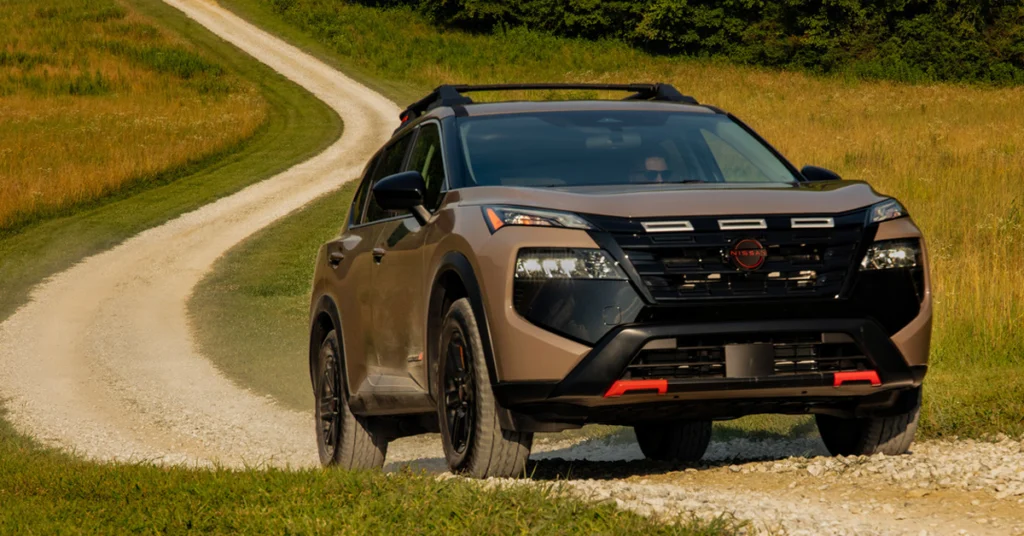 A 2026-Nissan-Rogue SUV navigates a winding gravel road through grassy fields, evoking an adventurous and serene mood. Trees line the background under a clear sky.