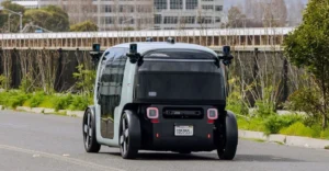 A rear-angle shot captures a two-tone, light gray and black autonomous vehicle on an asphalt road, with greenery and a building in the background.