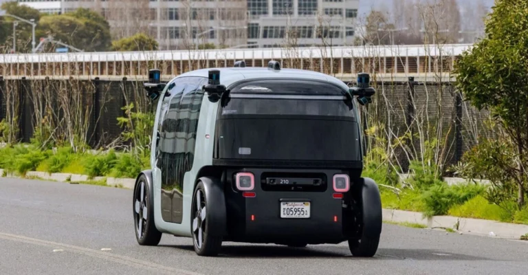 A rear-angle shot captures a two-tone, light gray and black autonomous vehicle on an asphalt road, with greenery and a building in the background.