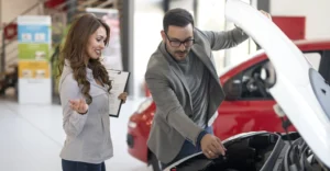 A medium shot captures a light-skinned woman and a light-skinned man in what appears to be a car dealership