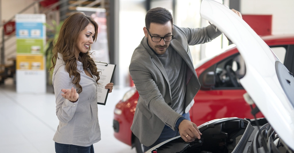 A medium shot captures a light-skinned woman and a light-skinned man in what appears to be a car dealership