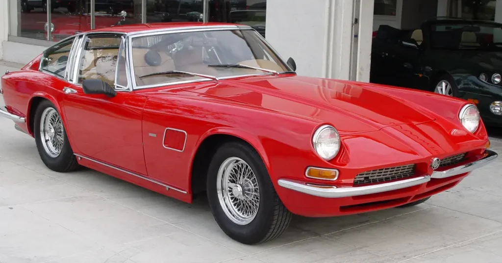 A red AC 428 Convertible (1966) sports car parked in front of a modern building, showcasing its sleek design and vibrant color.