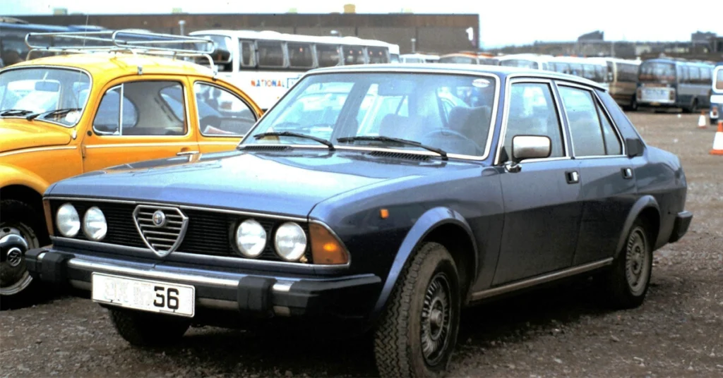 Blue vintage Alfa Romeo Alfa 6 (1979) sedan parked on gravel, with a yellow classic car beside it. Background shows several buses, creating a nostalgic, retro vibe.