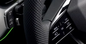 Close-up shot of a car's steering wheel, focusing on the left and right control panels. The steering wheel is covered in black perforated leather, providing a textured grip