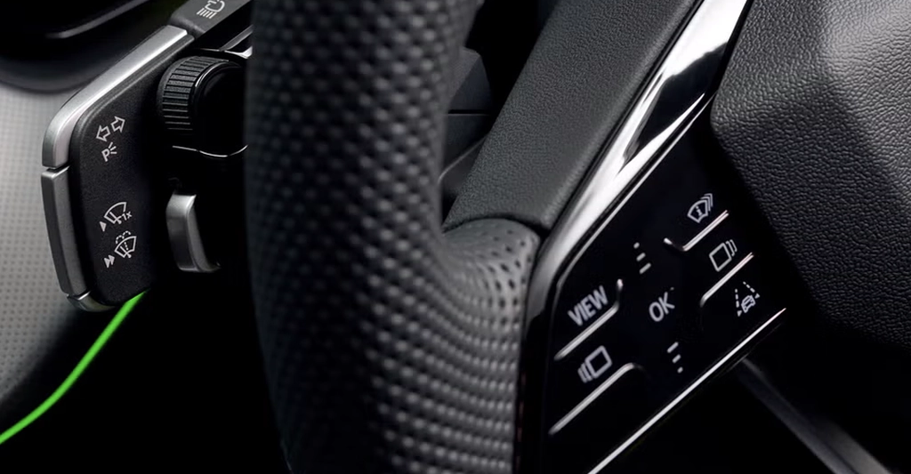 Close-up shot of a car's steering wheel, focusing on the left and right control panels. The steering wheel is covered in black perforated leather, providing a textured grip