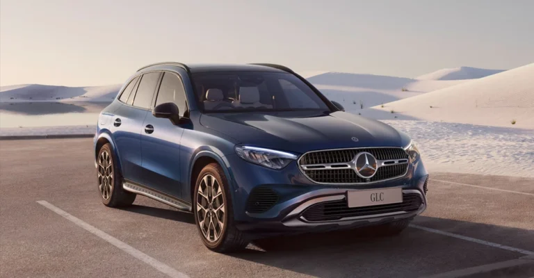 A blue Mercedes-Benz GLC SUV is parked on an asphalt surface with parking lines, set against a backdrop of white sand dunes under a clear sky.