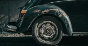 Close-up shot of the rear wheel and fender of a dark green vintage Volkswagen Beetle, showing signs of wear and rust, the paint is peeling off the fender, revealing patches of rust underneath