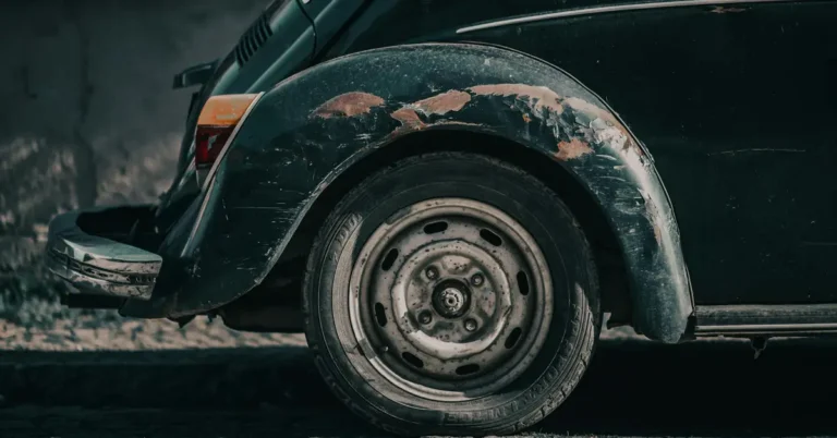 Close-up shot of the rear wheel and fender of a dark green vintage Volkswagen Beetle, showing signs of wear and rust, the paint is peeling off the fender, revealing patches of rust underneath