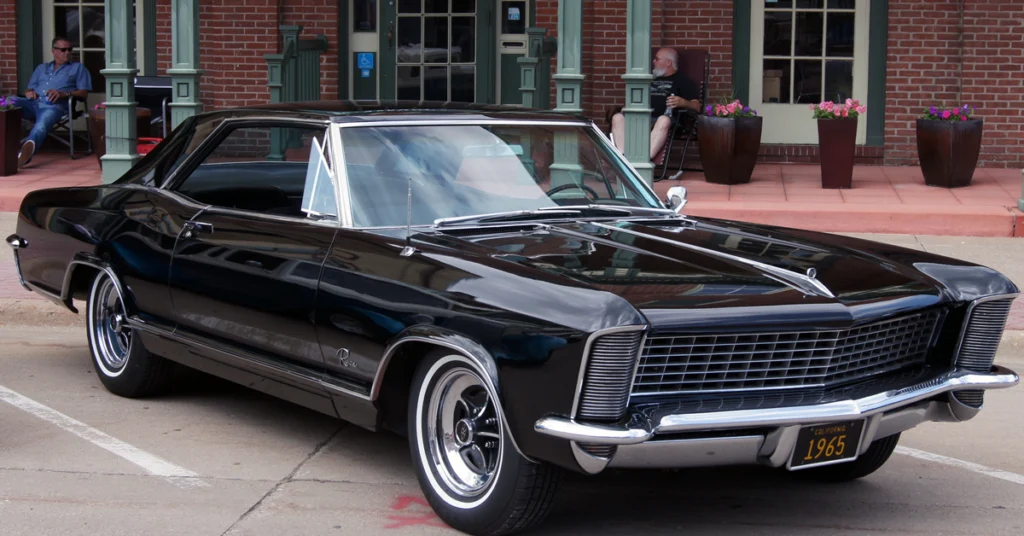 A sleek, black 1965 Buick Riviera is parked on a street. Its polished chrome accents and classic design exude elegance and nostalgia. Two people sit nearby against a brick building, creating a relaxed urban atmosphere.