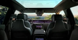 A view from the back seats of a Cadillac Escalade, looking forward towards the dashboard and windshield, with a large sunroof overhead. The car's interior is luxurious, featuring black leather seats with white stitching.