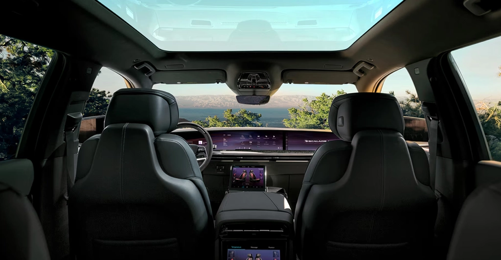 A view from the back seats of a Cadillac Escalade, looking forward towards the dashboard and windshield, with a large sunroof overhead. The car's interior is luxurious, featuring black leather seats with white stitching.