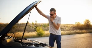 A medium shot captures a White man, around 30 years old, with short brown hair and wearing sunglasses, standing next to a car with its hood open.
