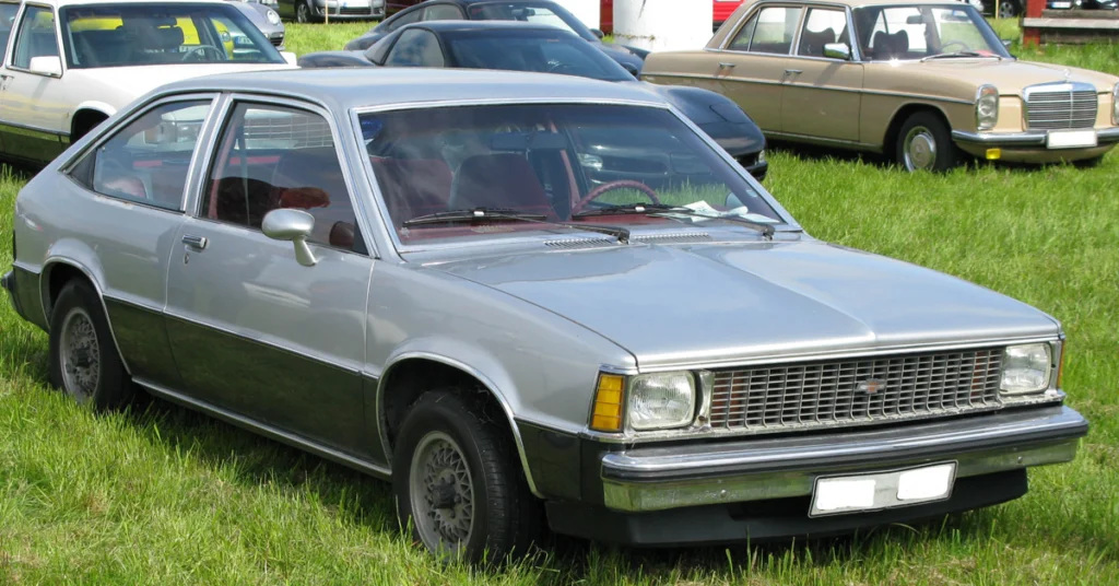 A vintage silver hatchback Chevrolet Citation X-11 (1980) car is parked on a grassy field, surrounded by other classic cars. The scene conveys a nostalgic, retro vibe.