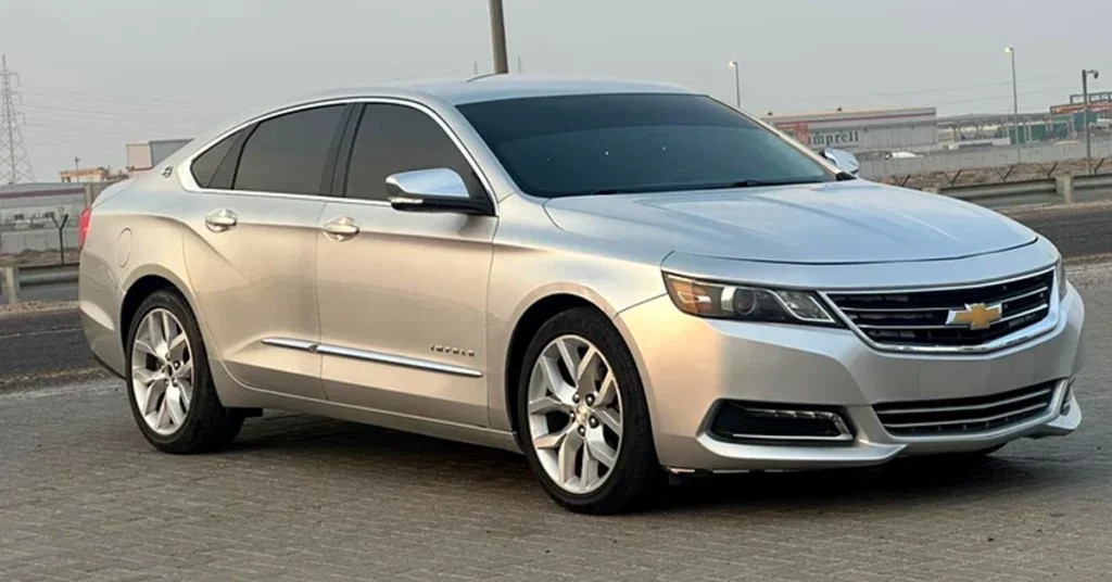 A silver Chevrolet-Impala sedan is parked on a paved surface. It has a sleek, modern design with tinted windows and the Chevrolet logo on the front grille. The background shows an industrial area under a cloudy sky.