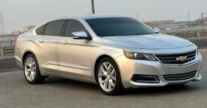 A silver Chevrolet-Impala sedan is parked on a paved surface. It has a sleek, modern design with tinted windows and the Chevrolet logo on the front grille. The background shows an industrial area under a cloudy sky.