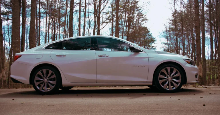 A white Chevrolet-Malibu sedan is parked on a concrete surface in a serene forest setting, surrounded by tall, leafless trees on a clear day, conveying tranquility.