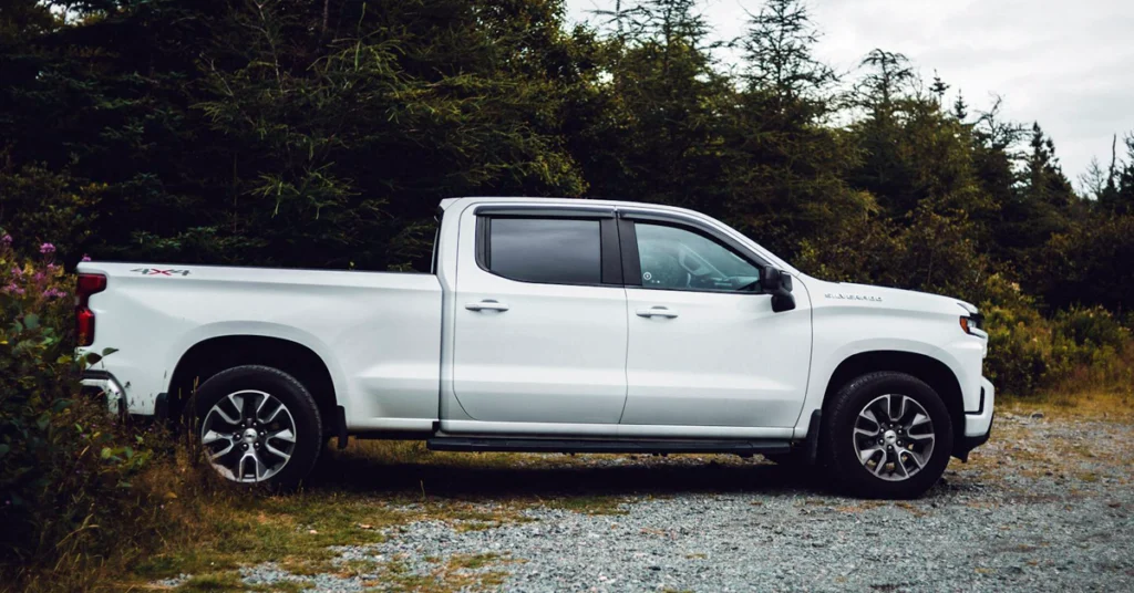 A white Chevrolet-Silverado-1500 pickup truck is parked on a gravel path next to dense green trees. The setting appears calm and natural, suggesting a rural or forest area.