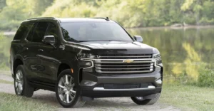 A dark gray Chevrolet Tahoe SUV is parked on a grassy area near a body of water. The SUV is facing forward, with its front end prominently displayed