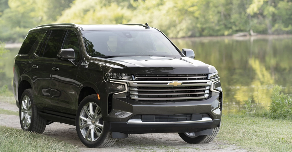 A dark gray Chevrolet Tahoe SUV is parked on a grassy area near a body of water. The SUV is facing forward, with its front end prominently displayed