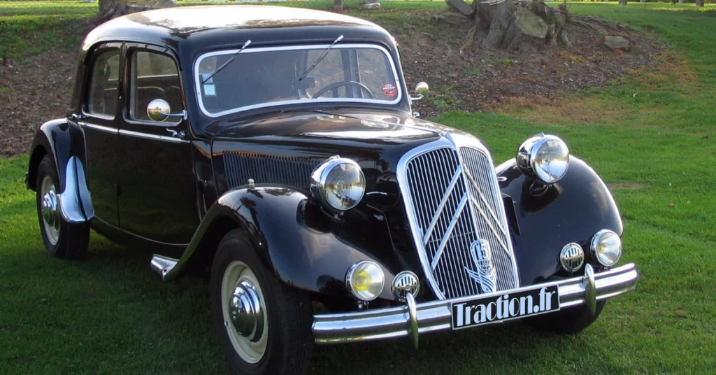 A vintage black Citroën Traction Avant (1934) car with shiny chrome accents and a distinctive grille is parked on green grass, with trees and rocks in the background, conveying classic elegance.