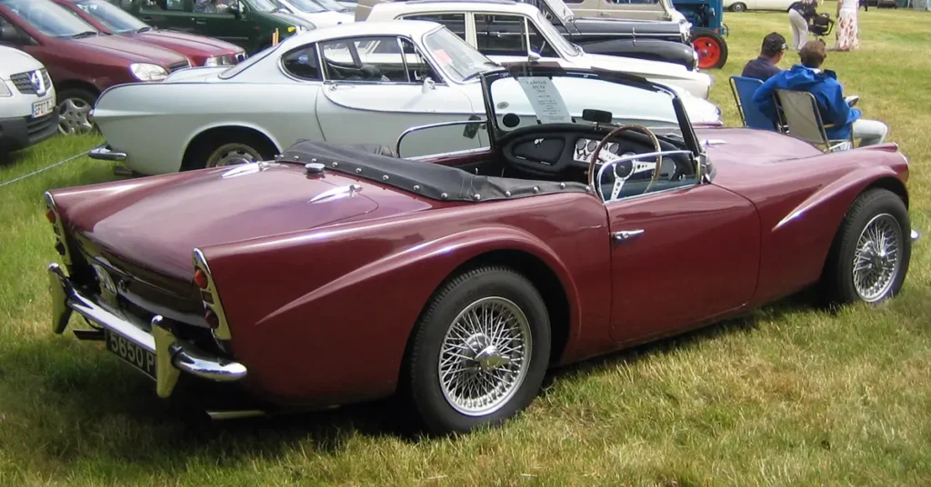 A classic car Daimler SP250 (1959) parked on grass beside several other cars in a casual outdoor setting.