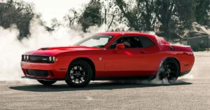 A red Dodge-Charger muscle car performing a burnout, releasing smoke from its rear tires on a sunlit road, surrounded by trees. The scene conveys power and excitement.