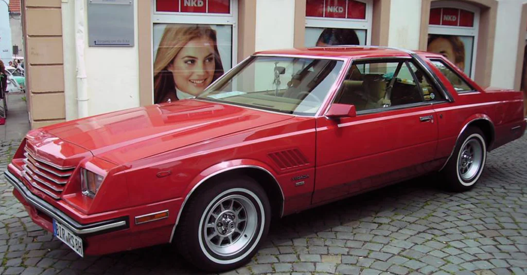 A shiny red vintage Dodge Mirada (1980) car parked on a cobblestone street. The car has a sleek design with chrome details. A smiling woman's poster is visible in the window behind.