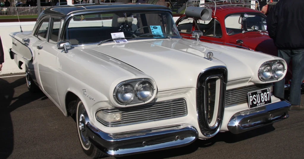 A white vintage Edsel (1958) car, likely from the 1950s, with distinctive circular headlights and chrome details, is parked at a car show. A red car is visible beside it.