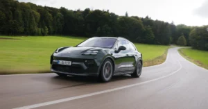 A sleek, dark green EV-Porsche-Macan SUV driving along a winding rural road, surrounded by lush greenery and trees under an overcast sky, conveying a sense of adventure.